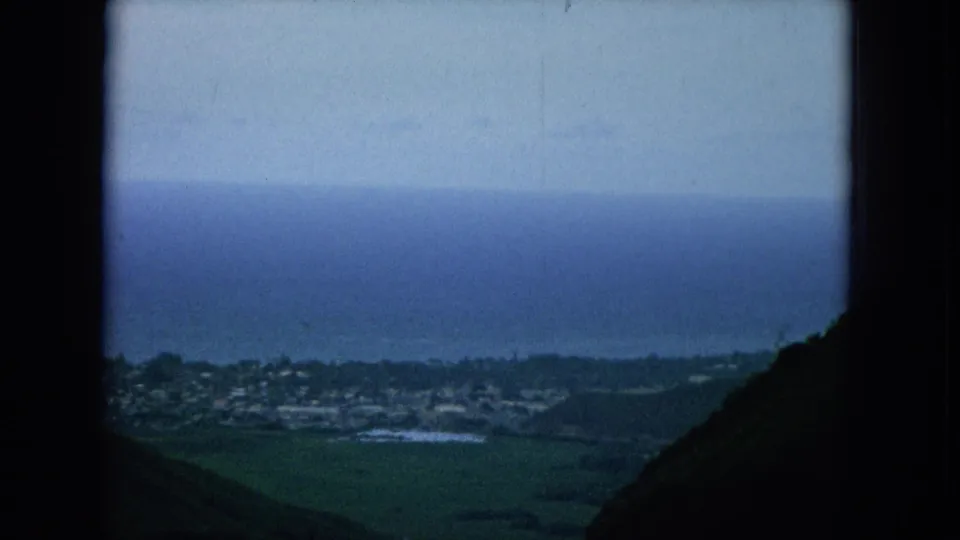 Elevated 1964 Oahu view across dark hills toward a coastal town, fields, and a blue ocean horizon.