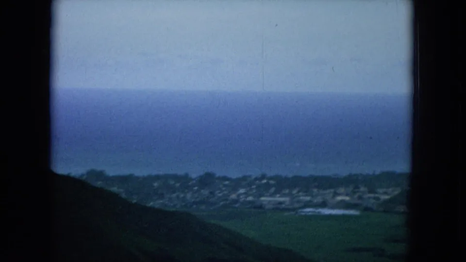 Handheld 8mm view of Oahu coastline with a distant town, green lowlands, dark slopes, and blue ocean beyond.