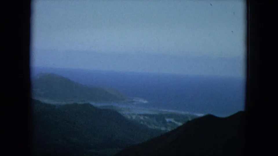 1964 Hawaii home-movie frame looks across dark hills toward a hazy shoreline, blue water, and a distant coastal headland.