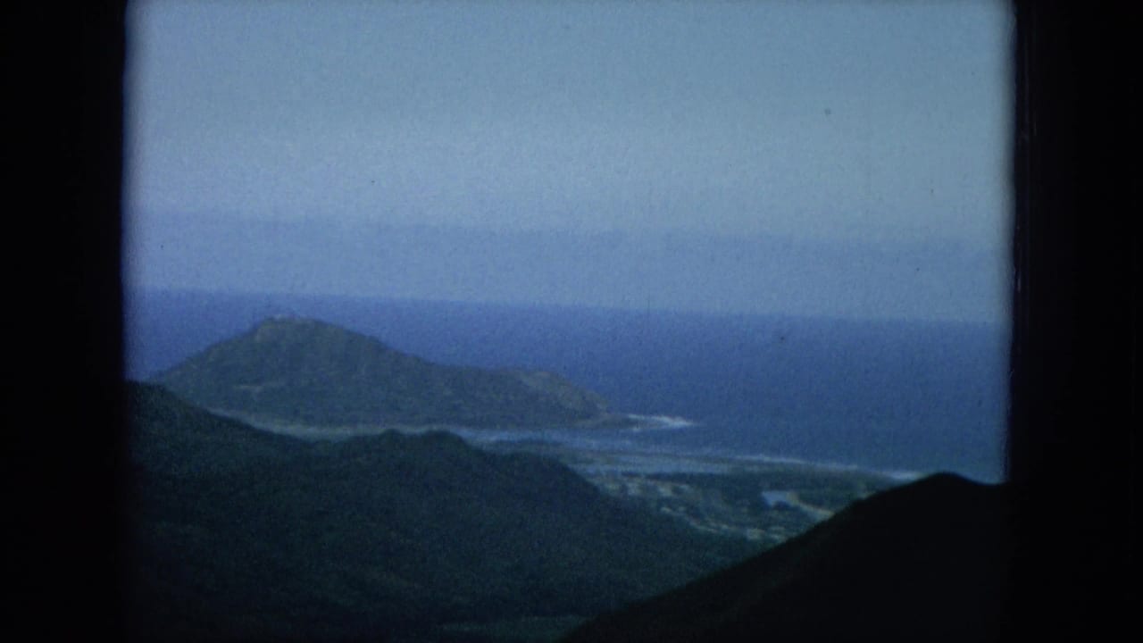 Elevated coastal view with dark film borders, mountain slopes, ocean, and a small offshore landform.