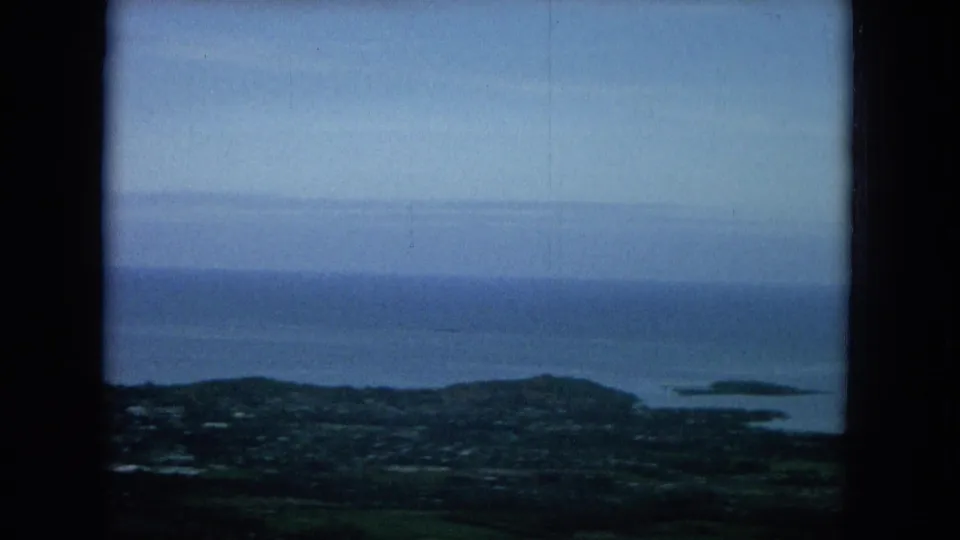 1964 Oahu view from above a coastal area, with the ocean horizon, low shoreline, and small offshore islands visible through dark framing.