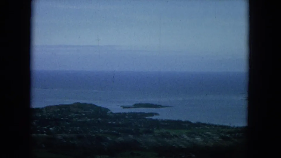 Elevated 1964 Oahu home-movie frame with a shoreline settlement, small offshore islands, green lowlands, and a blue ocean horizon.