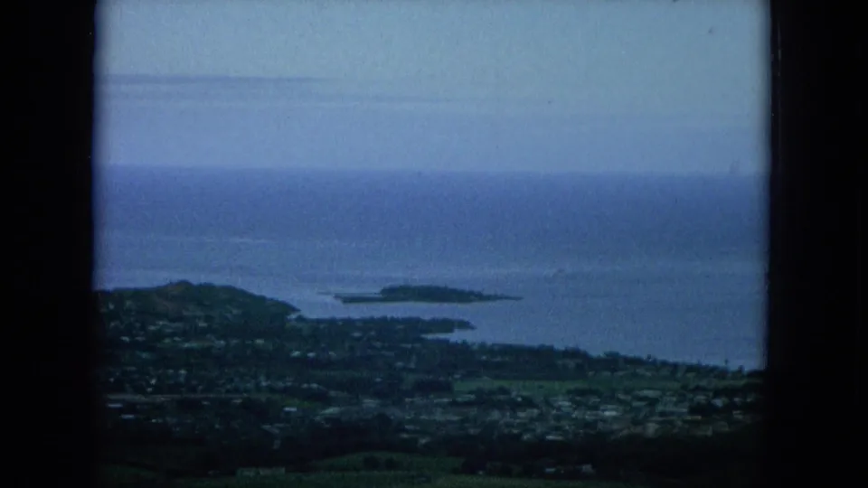 Final 1964 Oahu frame looking over green foreground and coastal development toward small islands and calm blue water.