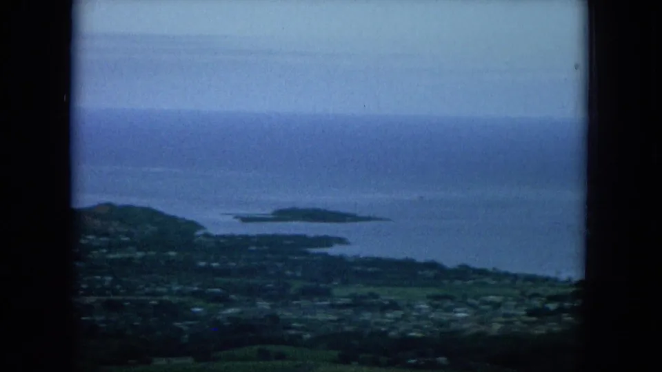 1964 Hawaii 8mm frame showing a high view of green coastal land, scattered settlement, blue water, and an offshore island shape.