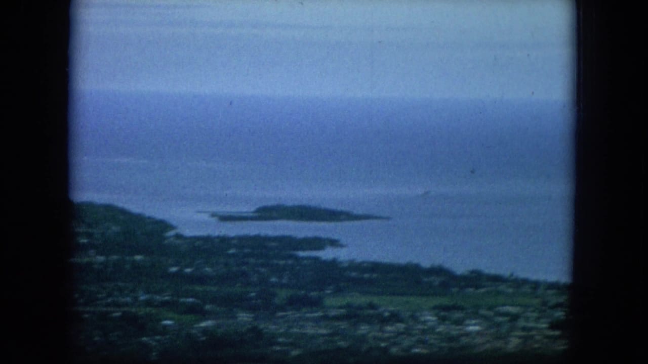 1964 Hawaii home-movie frame with coastal hills, town detail, calm blue ocean, and a low offshore island in the distance.