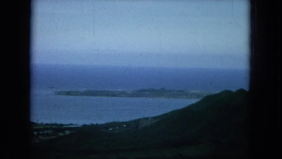 1964 Hawaii 8mm frame looking past a dark green hillside toward shoreline, open water, and a long low offshore island.