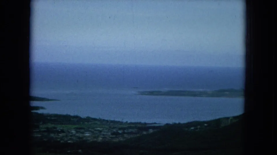 1964 Hawaii home-movie frame showing a wide elevated look over coastal settlement, dark hills, blue water, and distant island landforms.