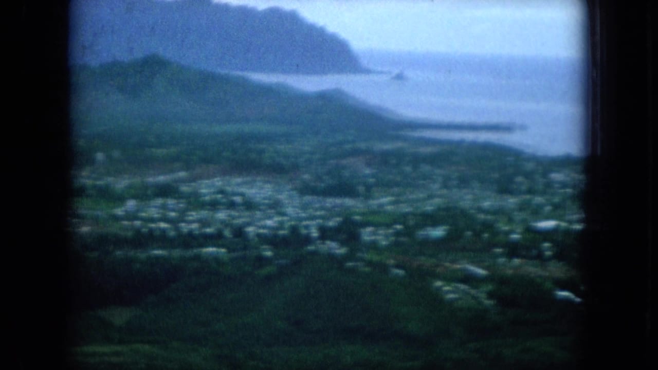 1964 8mm view over green Oahu hills, scattered homes, shoreline water, and distant coastal cliffs under a pale sky.
