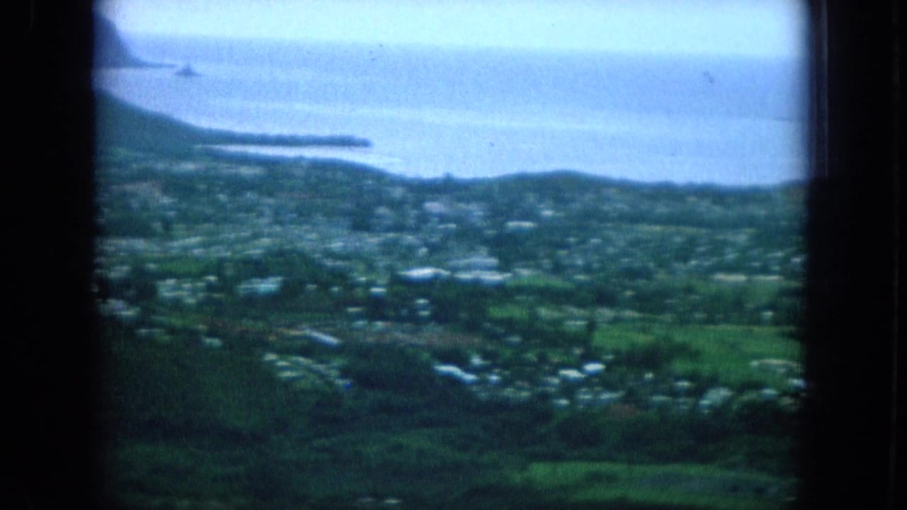 1964 Oahu home-movie frame showing green coastal land, small homes, blue ocean water, and low headlands in soft focus.
