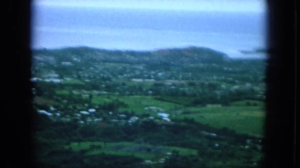 Soft 8mm Oahu view of green fields, clustered houses, low hills, and blue ocean water in the distance.