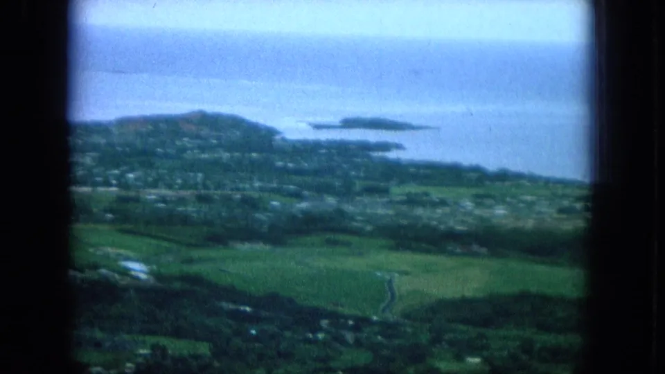 1964 Oahu frame with green fields, coastal neighborhoods, blue ocean, and small offshore landforms seen through soft 8mm grain.