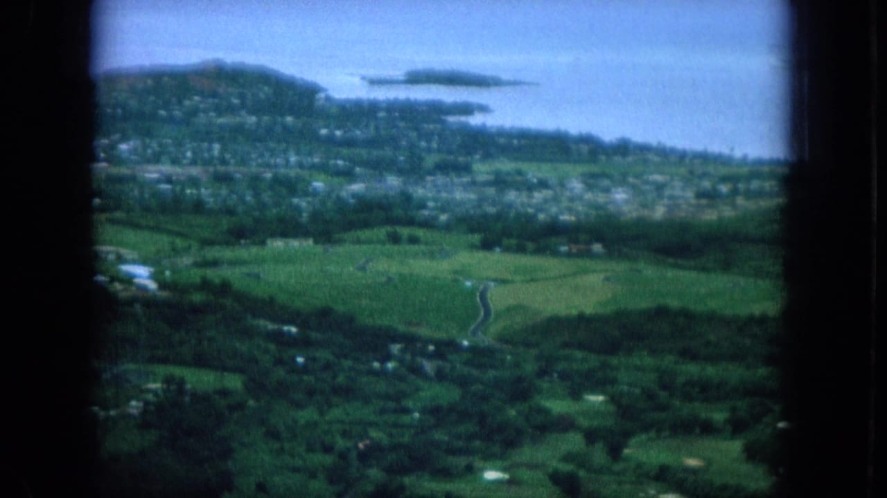 Aerial view of green hills, scattered buildings, shoreline water, and small islands through a dark window frame.
