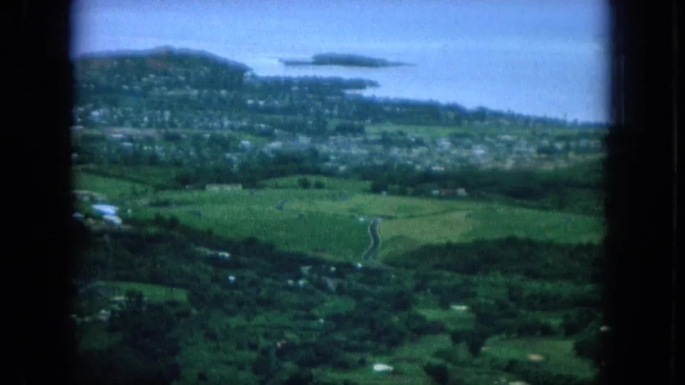 1964 Hawaii 8mm aerial view over green hills toward a coastal town, shoreline water, and distant small islands, framed by aircraft window edges.