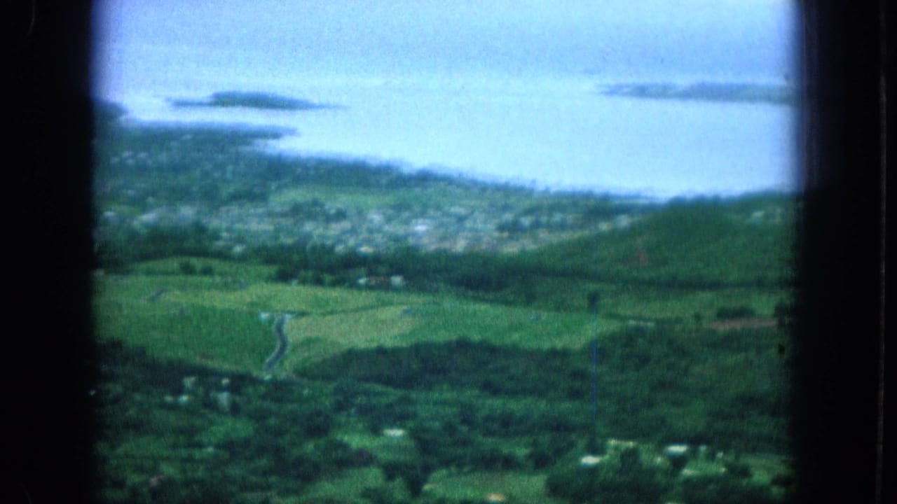 Aerial view over bright green fields and a coastal town, with blue water and small islands beyond.