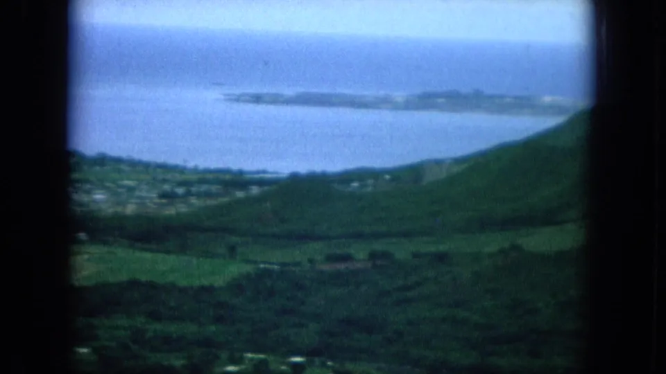 1964 Hawaii 8mm frame with soft aerial view of green hills dropping toward coastal water and a long low island offshore.