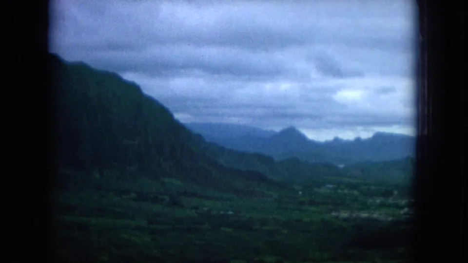 1964 Hawaii 8mm aerial view of a steep green mountain valley, cloudy sky, distant ridgelines, and lowland fields below.