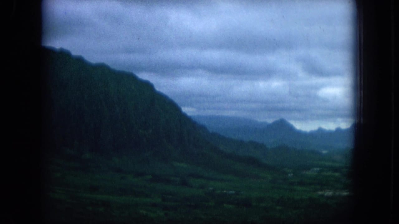 1964 Oahu home-movie view of misty green ridges and a valley through a dark window frame under a blue-gray overcast sky.