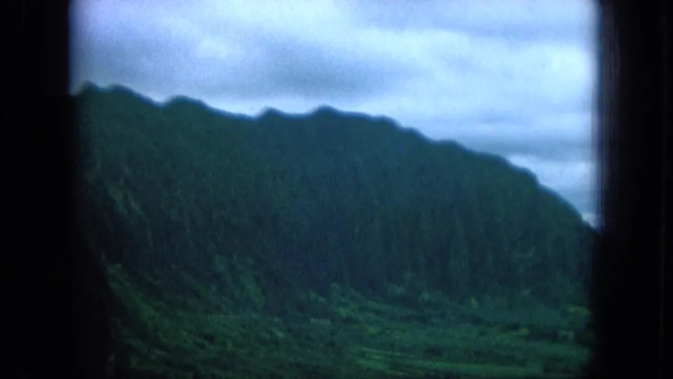 1964 Oahu home-movie frame of a dark green mountain slope above a lush valley, softened by overcast light and 8mm texture.