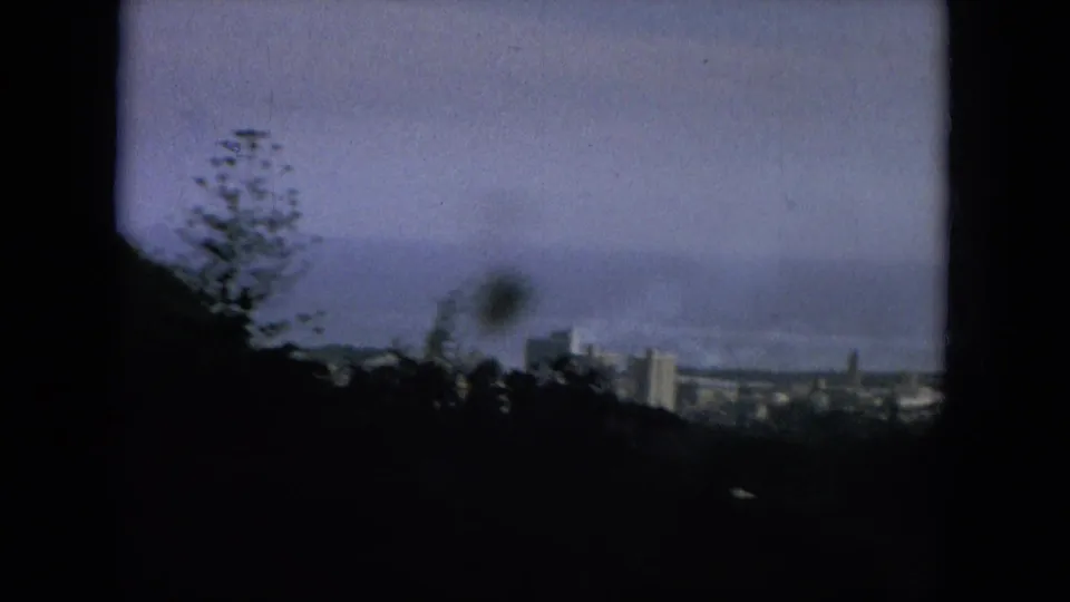 1964 Hawaii 8mm view over dark hillside vegetation toward distant high-rises, coastline, and hazy sky.