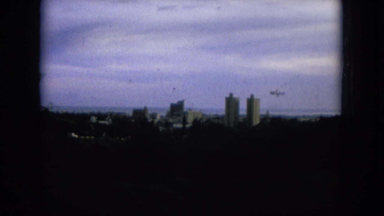 1964 8mm view of a distant Oahi, Hawaii skyline under a heavy overcast sky, with towers silhouetted above a dark foreground.