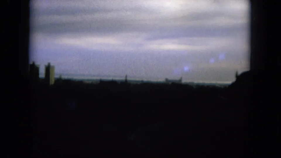 1964 home-movie frame showing a cloudy horizon and partial distant skyline, with the foreground falling into deep shadow.