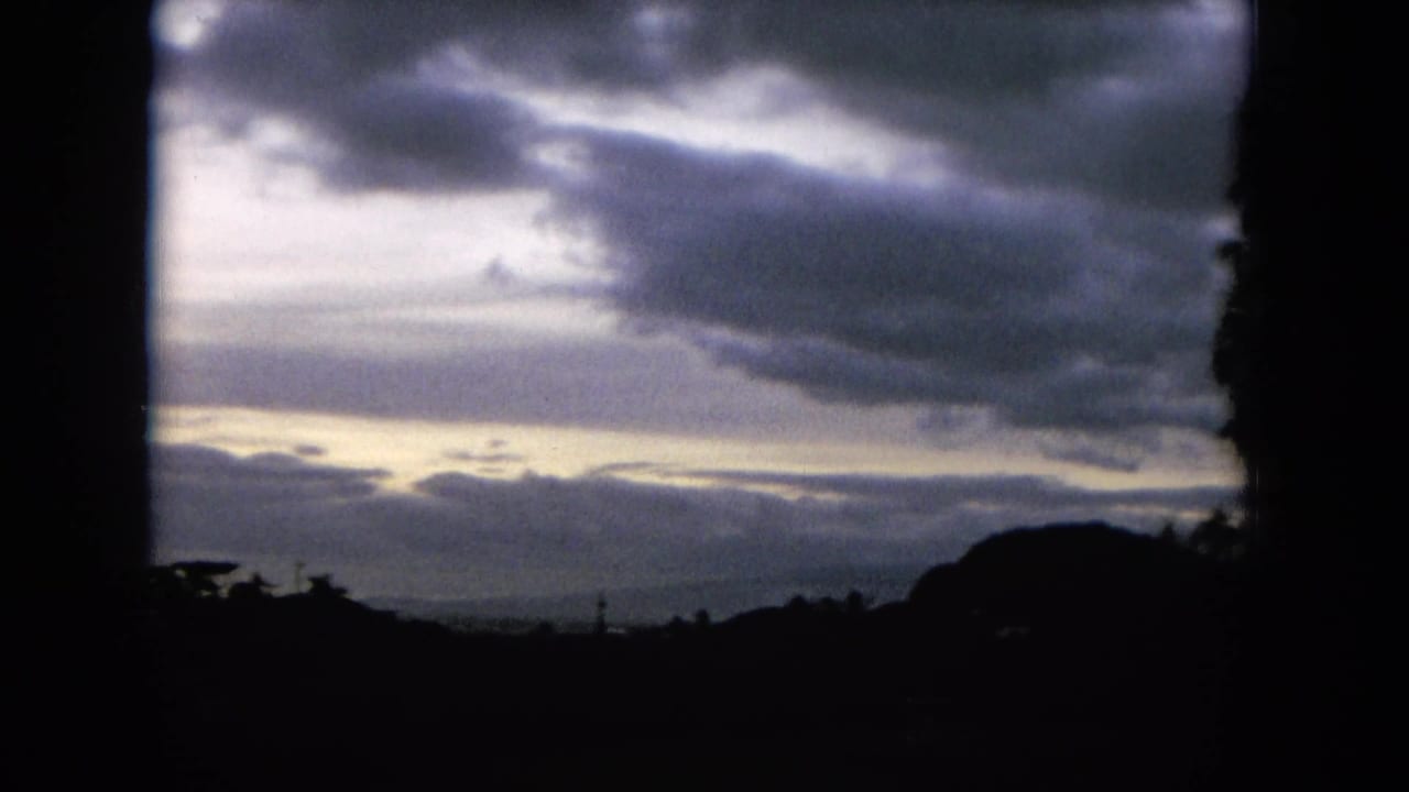 1964 Hawaii 8mm frame showing a cloud-heavy sky above dark hill and tree silhouettes, with pale light breaking near the horizon.