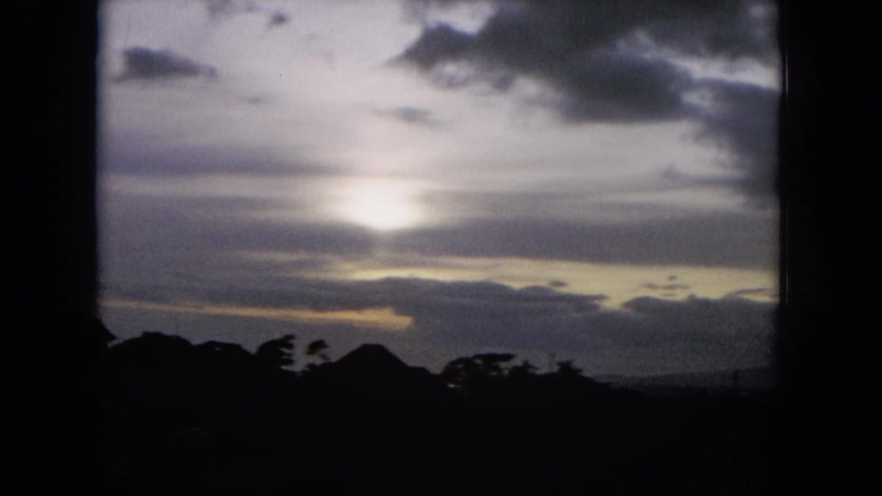 1964 Hawaii 8mm frame with a bright sun or moon diffused through layered clouds over a dark, tree-lined ridge.