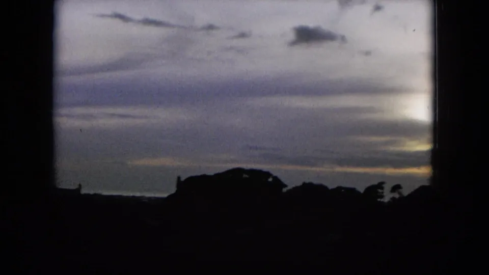 1964 Hawaii 8mm frame showing a muted cloud bank and bright edge of sun or moonlight above dark hill and tree silhouettes.