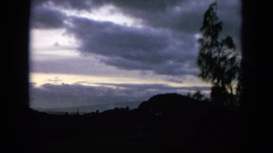 1964 Hawaii 8mm frame with heavy clouds, a bright horizon break, dark hills, and a silhouetted tree at the right edge.