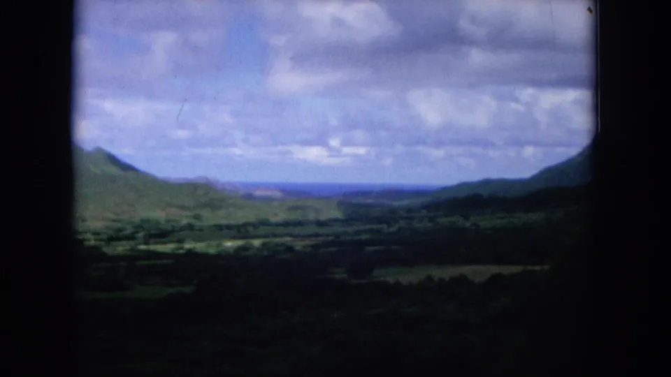 1964 Oahi, Hawaii 8mm frame looking across a green valley toward distant mountain slopes under a partly cloudy sky.