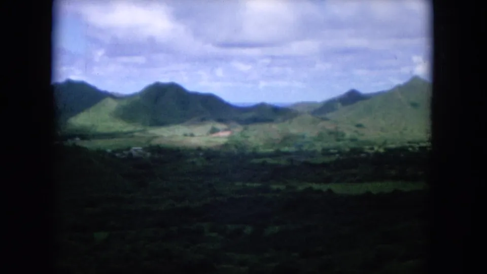 1964 Oahu home-movie view of green mountain ridges and a broad valley beneath a partly cloudy sky, framed by dark film edges.