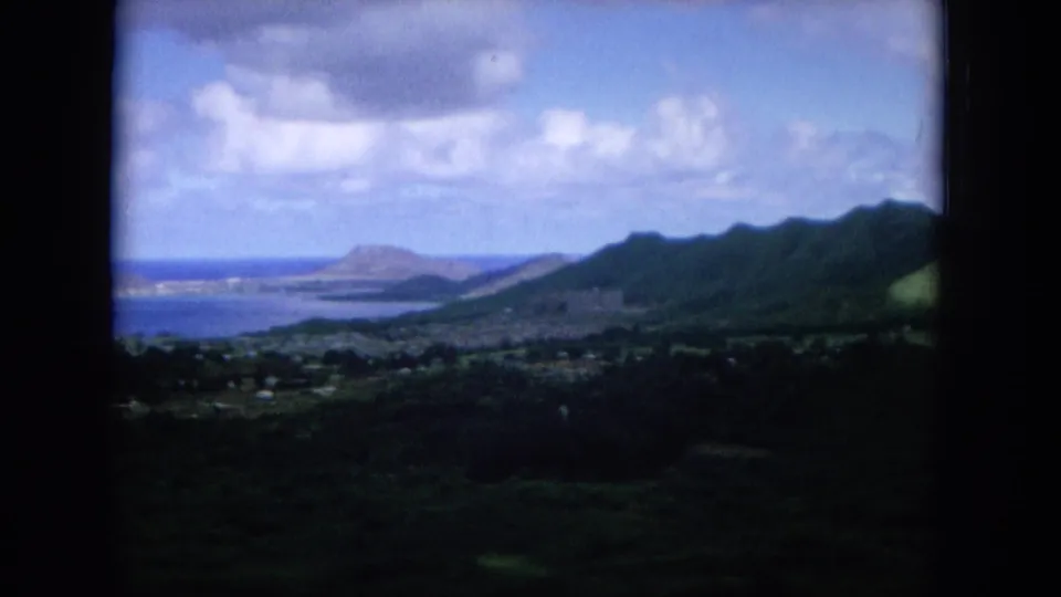 1964 Oahu home-movie frame showing green hills in the foreground, a coastal bay or ocean beyond, and a distant offshore landform.