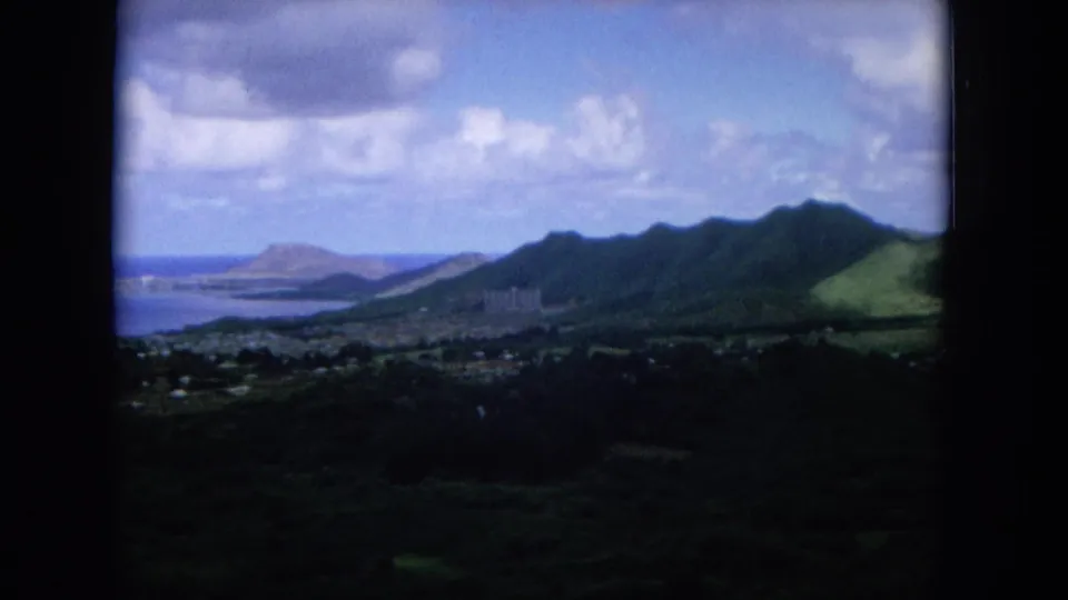 1964 Oahu elevated view across a green mountain ridge toward coastal water and a distant landform, with soft 8mm color and dark borders.