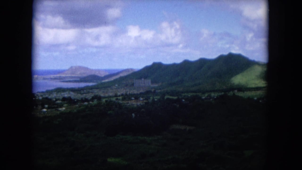 Aerial view of green mountains above a coastal town and bay under scattered clouds.