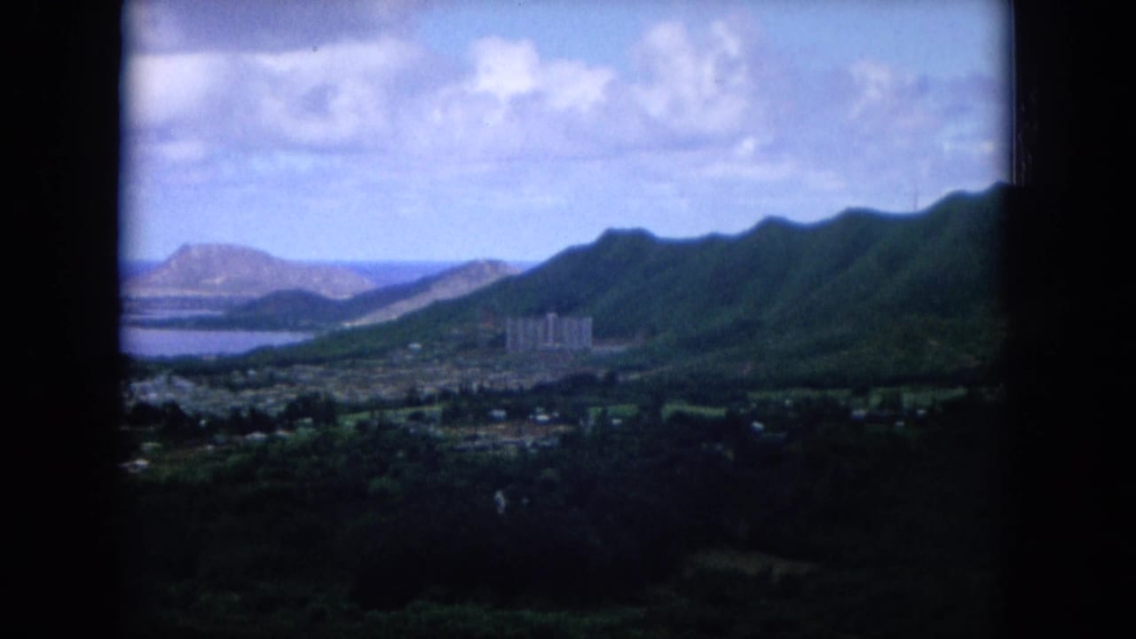 Oahu aerial landscape from 1964 footage, showing a coastal town, lush ridge, bay water, and distant shoreline.
