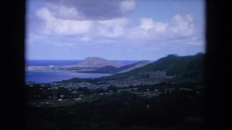 1964 Oahu aerial frame with a bay, distant volcanic-looking headland, coastal town, and green mountain slope.