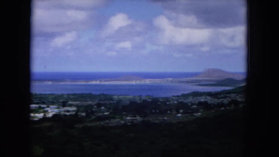 1964 Hawaii home-movie aerial view across an Oahu coastal town toward bay water, ocean, and a distant headland.