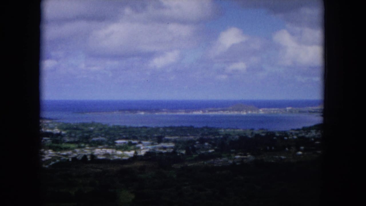 1964 8mm view from a high hill on Oahu, showing a coastal settlement, bay water, distant shoreline, and clouds.