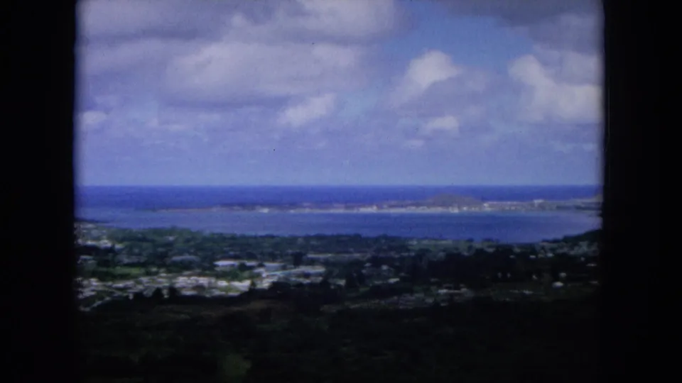 1964 8mm view from a high hill on Oahu, showing a coastal settlement, bay water, distant shoreline, and clouds.