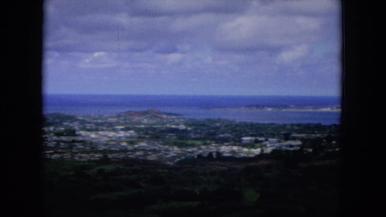 1964 Oahu panorama showing low-rise settlement below a hill, the Pacific beyond, and a broad cloud layer overhead.