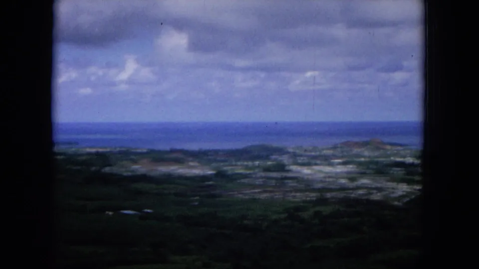 1964 8mm frame from an Oahu hillside, with green terrain, scattered light rooftops, and the ocean in the distance.
