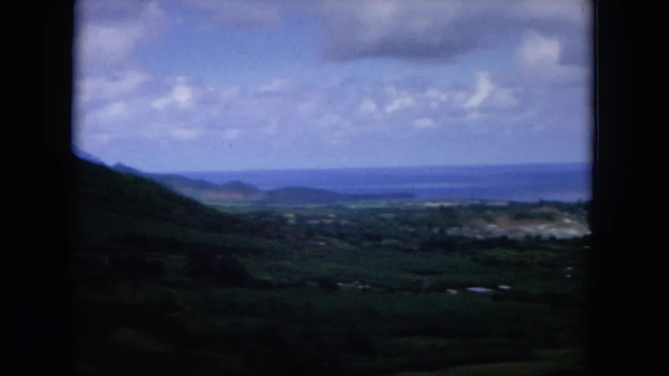 1964 Oahu hilltop panorama with a dark slope at left, coastal lowlands, distant blue water, and cloud cover.