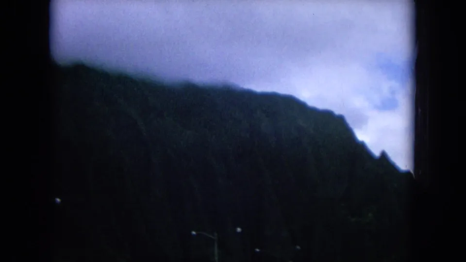 Shadowed 8mm view of an Oahu mountain slope from a moving vehicle, with soft focus and a cloudy sky.