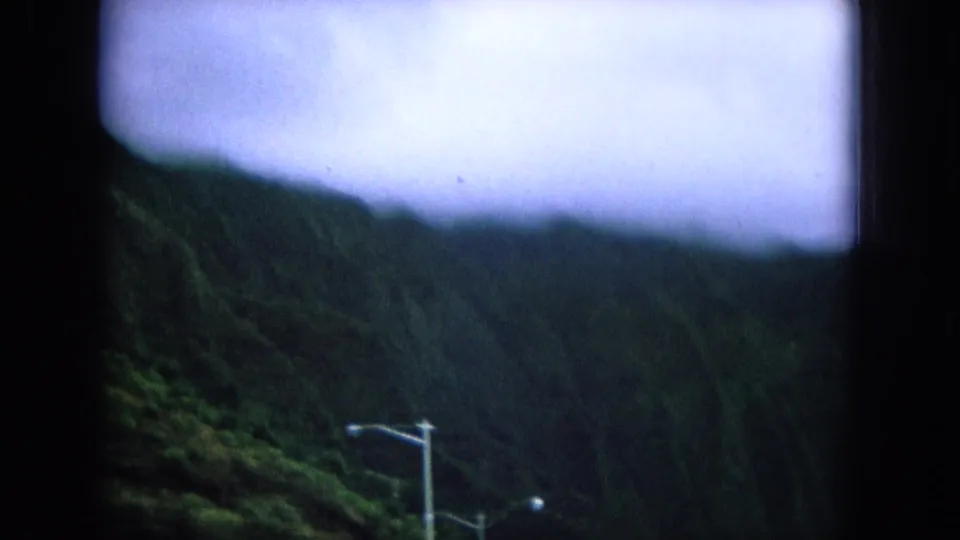 1964 Oahu roadside mountain view through a vehicle window, with green slopes, lamp posts, and overcast light.