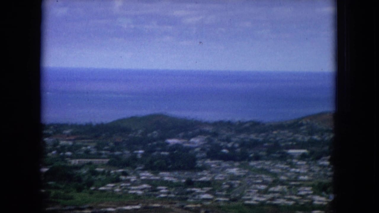 1964 Hawaii frame looking across a valley settlement toward dark green hills and the blue Pacific horizon.