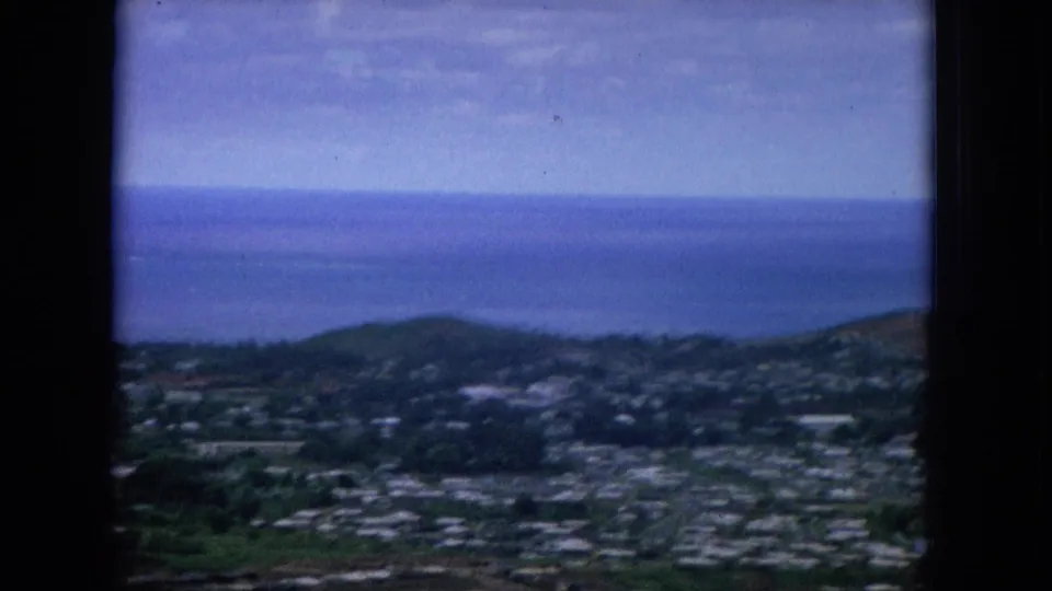 1964 Hawaii frame looking across a valley settlement toward dark green hills and the blue Pacific horizon.