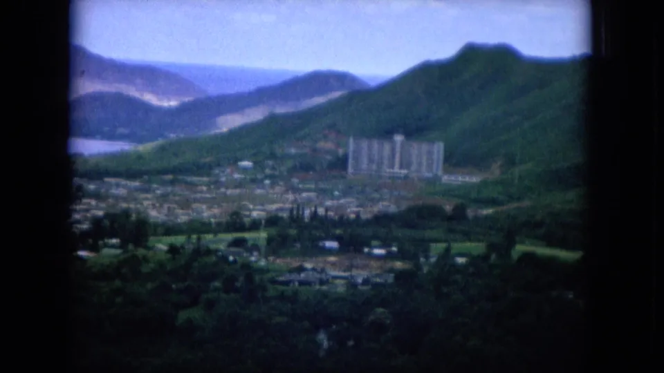 1964 Hawaii 8mm frame showing a valley settlement, water inlet, steep green slopes, and a prominent multi-story building.