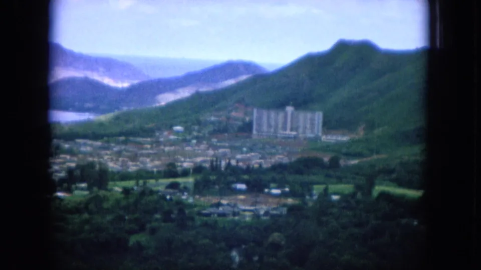 1964 Hawaii home-movie frame with a valley town, prominent building, forested slopes, and blue water beyond.