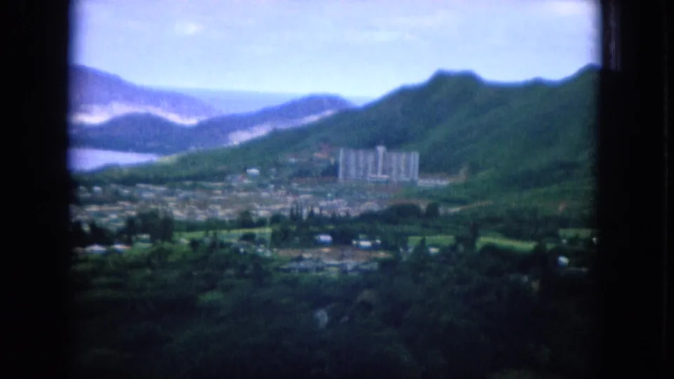 1964 Hawaii home-movie frame showing green ridges, a coastal town, distant water, and tall buildings from a hillside viewpoint.