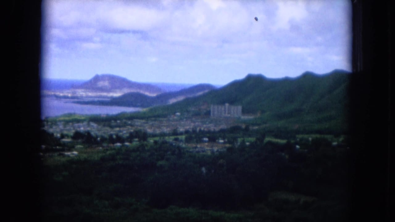 Wider coastal panorama with mountains, ocean, town blocks, and distant headland under a cloudy sky.